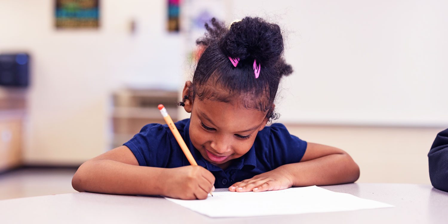 Young student smiling while writing with a pencil at a desk.