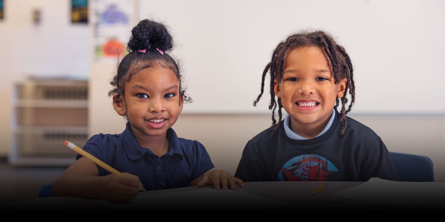 Two smiling students at a classroom desk, one holding a pencil.